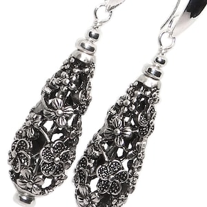 May include: A pair of silver-toned dangle earrings. Each earring features a teardrop-shaped design, intricately detailed with floral patterns. The earrings have a hook closure and are set against a white background.