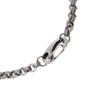 May include: A silver-toned metal chain necklace featuring round, textured links with an intricate, repeating pattern. The chain is secured with a smooth, elongated lobster claw clasp that connects to the patterned links on both ends of the jewelry piece.