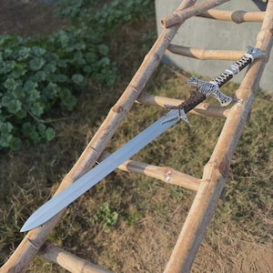 May include: A double-edged sword with a long, pointed steel blade. The hilt features a dark brown, textured grip wrapped in white cord, accented by an ornate, silver-colored crossguard with intricate scrollwork and a matching pommel.