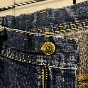 May include: Close-up of a pair of blue denim jeans. The jeans feature a brass button with the brand name "Wrangler" and a zipper. Yellow stitching accents the seams.