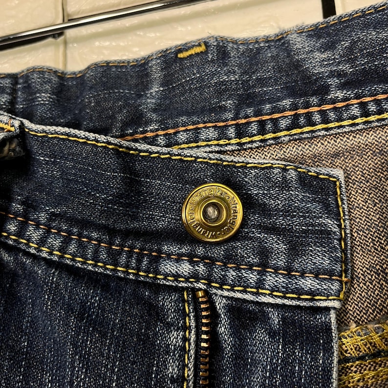 May include: Close-up of a pair of blue denim jeans. The jeans feature a brass button with the brand name "Wrangler" and a zipper. Yellow stitching accents the seams.