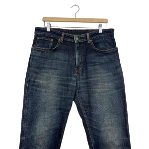 Puede incluir: Vaqueros de mezclilla de lavado oscuro colgados de una percha de madera. Los vaqueros tienen un dise&ntilde;o cl&aacute;sico de cinco bolsillos con cierre de bot&oacute;n y cremallera. La mezclilla tiene un aspecto descolorido y desgastado.