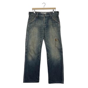 May include: Faded blue denim jeans with a distressed, worn appearance. The jeans have a relaxed fit, a button and zip fastening, and a single ripped area on the right leg. The jeans are hanging on a wooden hanger.