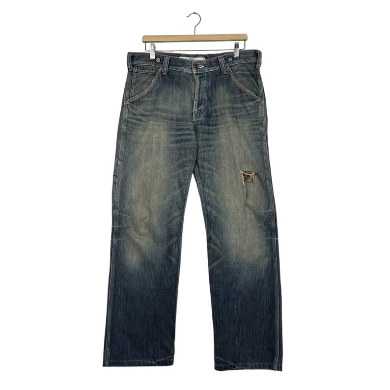May include: Faded blue denim jeans with a distressed, worn appearance. The jeans have a relaxed fit, a button and zip fastening, and a single ripped area on the right leg. The jeans are hanging on a wooden hanger.