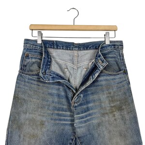 K&ouml;nnte beinhalten: Blaue Jeans mit verblichener Waschung und sichtbaren Gebrauchsspuren. Die Jeans h&auml;ngen an einem Holzkleiderb&uuml;gel, der Rei&szlig;verschluss ist teilweise ge&ouml;ffnet. Der Stoff weist hellere Stellen und dunklere Flecken auf.