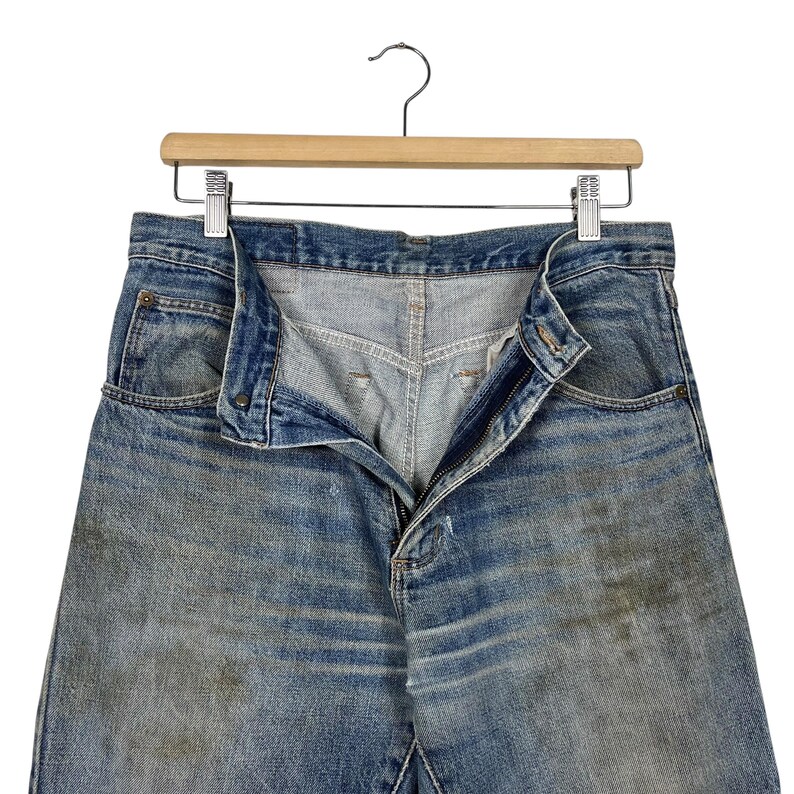 K&ouml;nnte beinhalten: Blaue Jeans mit verblichener Waschung und sichtbaren Gebrauchsspuren. Die Jeans h&auml;ngen an einem Holzkleiderb&uuml;gel, der Rei&szlig;verschluss ist teilweise ge&ouml;ffnet. Der Stoff weist hellere Stellen und dunklere Flecken auf.