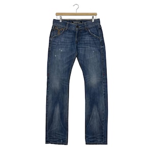 K&ouml;nnte beinhalten: Blaue Jeans mit verwaschener Optik, h&auml;ngen an einem Holzb&uuml;gel. Die Jeans haben ein klassisches F&uuml;nf-Taschen-Design mit Knopf- und Rei&szlig;verschluss. Es gibt hellfarbene Farbspritzer und ein dekoratives Design auf der Vordertasche.