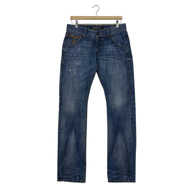 K&ouml;nnte beinhalten: Blaue Jeans mit verwaschener Optik, h&auml;ngen an einem Holzb&uuml;gel. Die Jeans haben ein klassisches F&uuml;nf-Taschen-Design mit Knopf- und Rei&szlig;verschluss. Es gibt hellfarbene Farbspritzer und ein dekoratives Design auf der Vordertasche.