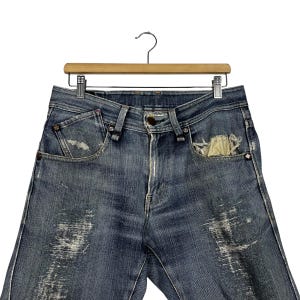 K&ouml;nnte beinhalten: Blaue Jeans aus Denim mit Used-Details, einschlie&szlig;lich verblichener Patches und einer zerrissenen Tasche. Die Jeans werden auf einem Holzb&uuml;gel pr&auml;sentiert, der die Vorderseite des Kleidungsst&uuml;cks zeigt. Die Jeans haben einen Knopfverschluss und ein klassisches F&uuml;nf-Taschen-Design.