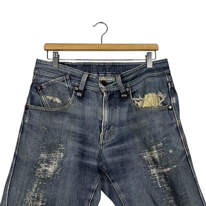 K&ouml;nnte beinhalten: Blaue Jeans aus Denim mit Used-Details, einschlie&szlig;lich verblichener Patches und einer zerrissenen Tasche. Die Jeans werden auf einem Holzb&uuml;gel pr&auml;sentiert, der die Vorderseite des Kleidungsst&uuml;cks zeigt. Die Jeans haben einen Knopfverschluss und ein klassisches F&uuml;nf-Taschen-Design.