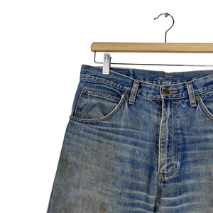 K&ouml;nnte beinhalten: Verblasste blaue Jeansshorts h&auml;ngen an einem Holzkleiderb&uuml;gel. Die Shorts haben ein klassisches F&uuml;nf-Taschen-Design, einen Knopfverschluss und G&uuml;rtelschlaufen. Der Denim weist Gebrauchsspuren und Ausbleichungen auf.