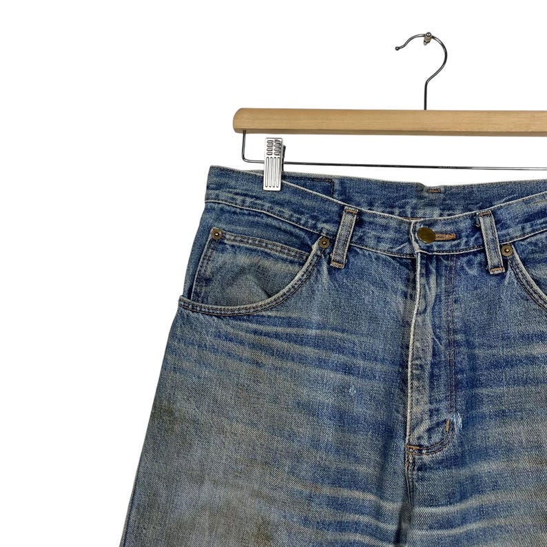 K&ouml;nnte beinhalten: Verblasste blaue Jeansshorts h&auml;ngen an einem Holzkleiderb&uuml;gel. Die Shorts haben ein klassisches F&uuml;nf-Taschen-Design, einen Knopfverschluss und G&uuml;rtelschlaufen. Der Denim weist Gebrauchsspuren und Ausbleichungen auf.