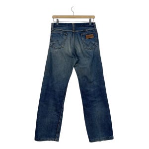 Puede incluir: Vaqueros azules de mezclilla colgados de una percha de madera. Los vaqueros tienen un dise&ntilde;o cl&aacute;sico de cinco bolsillos, un parche de cuero marr&oacute;n y el logotipo de Wrangler. Los vaqueros son de un lavado medio con algo de decoloraci&oacute;n.