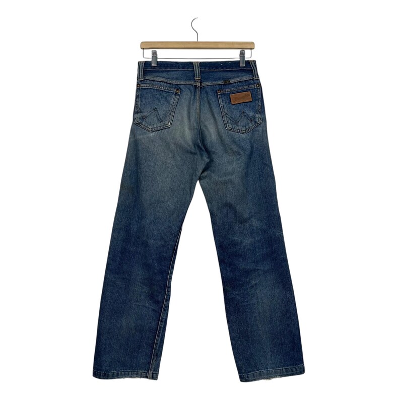 Puede incluir: Vaqueros azules de mezclilla colgados de una percha de madera. Los vaqueros tienen un dise&ntilde;o cl&aacute;sico de cinco bolsillos, un parche de cuero marr&oacute;n y el logotipo de Wrangler. Los vaqueros son de un lavado medio con algo de decoloraci&oacute;n.