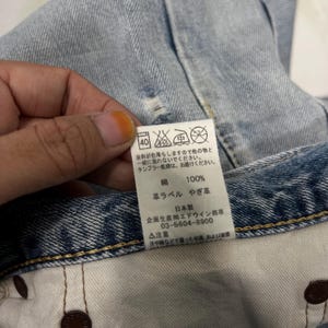 May include: Close-up of a light blue denim garment with a white tag. The tag features Japanese text and laundry symbols. The garment has a distressed look with visible wear and tear.