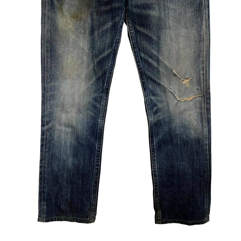 May include: Faded blue denim jeans with distressed details, including a tear on the right knee. The jeans have a classic five-pocket design and a straight leg cut. The denim shows signs of wear and fading.