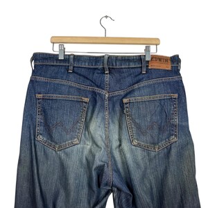 K&ouml;nnte beinhalten: Blaue Jeans aus Denim mit verwaschener Optik, mit zwei Ges&auml;&szlig;taschen und einem braunen Lederaufn&auml;her mit dem Markennamen "Edwin". Die Jeans h&auml;ngen an einem Holzb&uuml;gel vor wei&szlig;em Hintergrund.