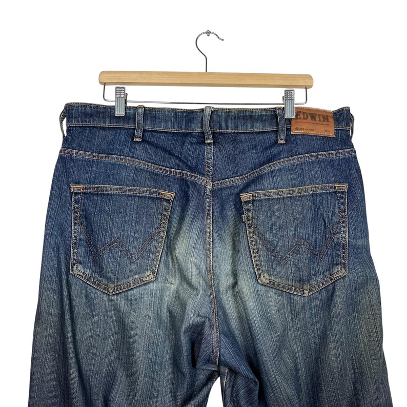 K&ouml;nnte beinhalten: Blaue Jeans aus Denim mit verwaschener Optik, mit zwei Ges&auml;&szlig;taschen und einem braunen Lederaufn&auml;her mit dem Markennamen "Edwin". Die Jeans h&auml;ngen an einem Holzb&uuml;gel vor wei&szlig;em Hintergrund.