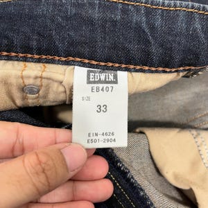 Puede incluir: Primer plano de unos vaqueros de mezclilla azul oscuro con una etiqueta de talla 33. La etiqueta dice "Edwin" e incluye el n&uacute;mero de modelo EB407. Informaci&oacute;n adicional en la etiqueta incluye "EIN-4626" y "E501-2904".