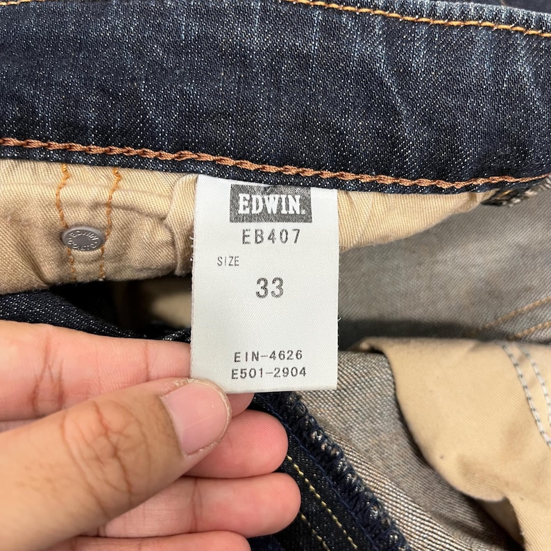 Puede incluir: Primer plano de unos vaqueros de mezclilla azul oscuro con una etiqueta de talla 33. La etiqueta dice "Edwin" e incluye el n&uacute;mero de modelo EB407. Informaci&oacute;n adicional en la etiqueta incluye "EIN-4626" y "E501-2904".