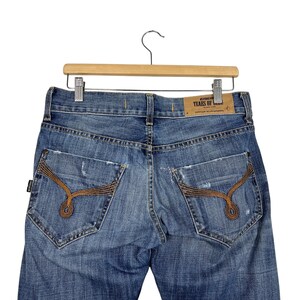 K&ouml;nnte beinhalten: Hellblaue Jeans mit Used-Details und einzigartigen braunen N&auml;hten auf den Ges&auml;&szlig;taschen. Die Jeans werden auf einem Holzb&uuml;gel pr&auml;sentiert. Ein Lederaufn&auml;her mit dem Text "TEARS OF BEAN" ist sichtbar.