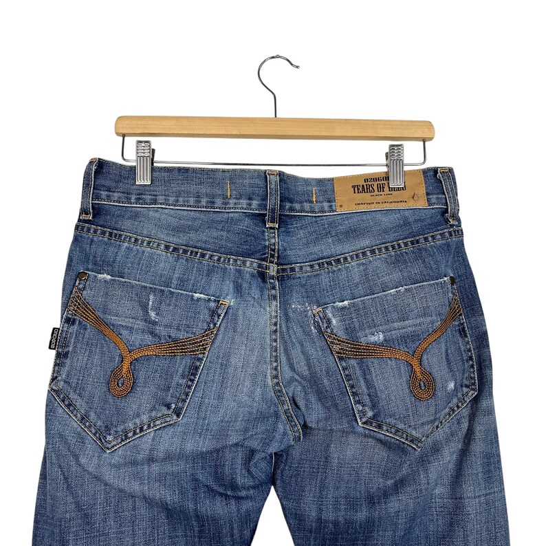 K&ouml;nnte beinhalten: Hellblaue Jeans mit Used-Details und einzigartigen braunen N&auml;hten auf den Ges&auml;&szlig;taschen. Die Jeans werden auf einem Holzb&uuml;gel pr&auml;sentiert. Ein Lederaufn&auml;her mit dem Text "TEARS OF BEAN" ist sichtbar.