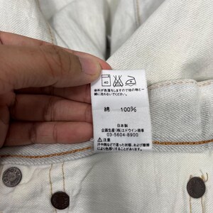 May include: Close-up of a white fabric tag with Japanese text and symbols. The tag indicates the fabric is 100% cotton and provides washing instructions. The tag also includes a phone number and a warning about color transfer.