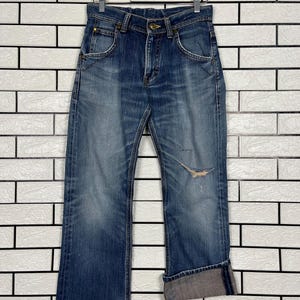 May include: Blue denim jeans with a distressed look, featuring a faded wash and a tear on the right leg. The jeans have a classic five-pocket design, a button and zipper closure, and are cuffed at the ankles.