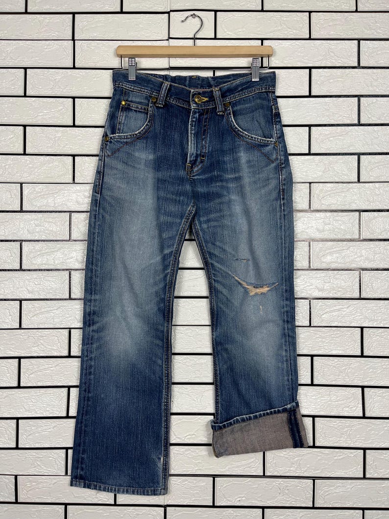 May include: Blue denim jeans with a distressed look, featuring a faded wash and a tear on the right leg. The jeans have a classic five-pocket design, a button and zipper closure, and are cuffed at the ankles.