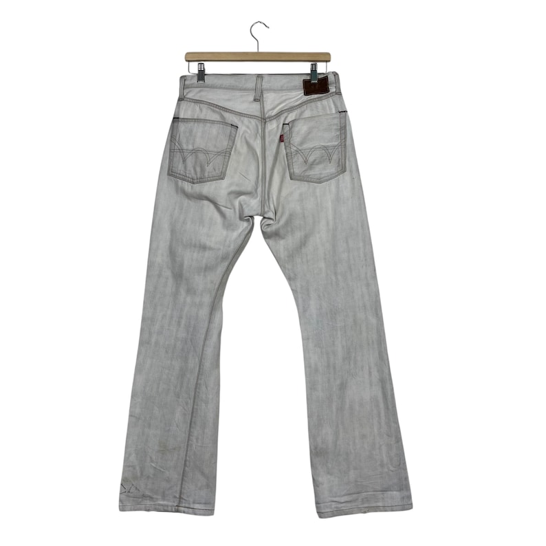 May include: Light gray denim jeans hanging on a wooden hanger. The jeans have a faded appearance with visible stitching and pockets. A small brown leather patch is visible on the back. The jeans are a classic style.