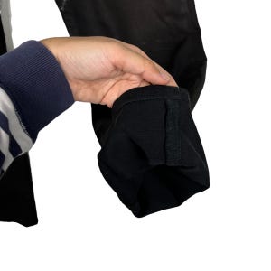 May include: Close-up of black denim pants. The cuff of the pants is folded up, revealing the hem. The pants are held by a person wearing a blue and white striped sleeve.