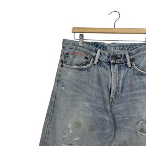 May include: Light blue denim jeans with distressed details, including faded patches and a torn knee. The jeans are hanging on a wooden hanger with a metal hook. The jeans have a classic five-pocket design.