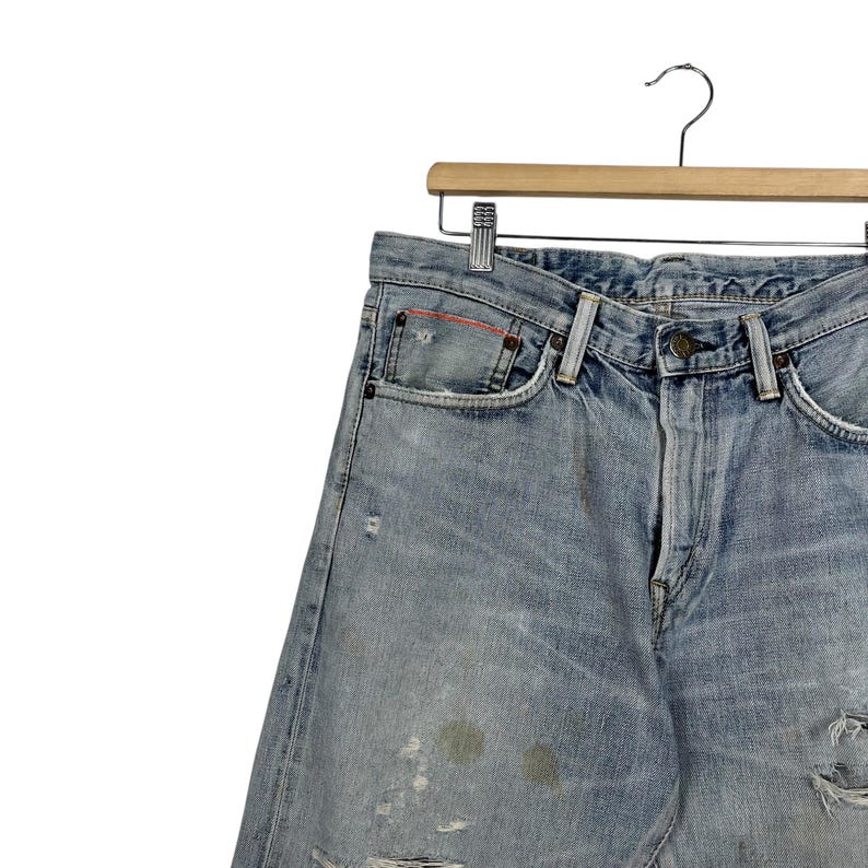 May include: Light blue denim jeans with distressed details, including faded patches and a torn knee. The jeans are hanging on a wooden hanger with a metal hook. The jeans have a classic five-pocket design.