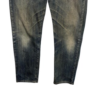 May include: Close-up of the lower portion of a pair of blue denim jeans. The jeans have a faded, worn appearance with lighter patches and subtle distressing. The fabric shows vertical and horizontal wear patterns.