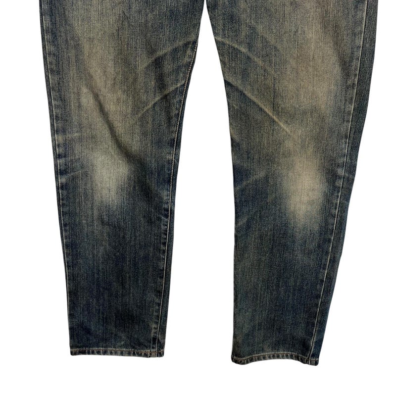 May include: Close-up of the lower portion of a pair of blue denim jeans. The jeans have a faded, worn appearance with lighter patches and subtle distressing. The fabric shows vertical and horizontal wear patterns.