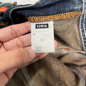 May include: Close-up of a denim garment tag. The tag reads "EDWIN" with the style number "405XV8" and size "32". The garment has orange stitching and a brown interior.