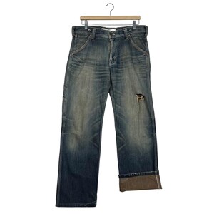May include: Blue denim jeans with a distressed appearance. The jeans have a relaxed fit, a button and zip closure, and a small rip on the right leg. The cuffs are rolled up, revealing a brown lining. The jeans are hanging on a wooden hanger.