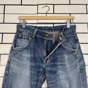 May include: Blue denim jeans with a button and zipper closure, hanging on a wooden hanger. The jeans have front pockets and a faded wash. The jeans are displayed against a white brick wall.