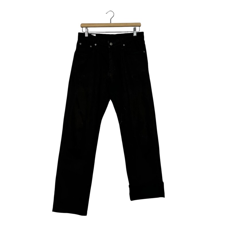 May include: Black denim jeans hanging on a wooden hanger. The jeans have a classic five-pocket design, a button and zipper closure, and a straight leg fit. The cuffs are rolled up, and the jeans are made of a sturdy, dark-colored denim.