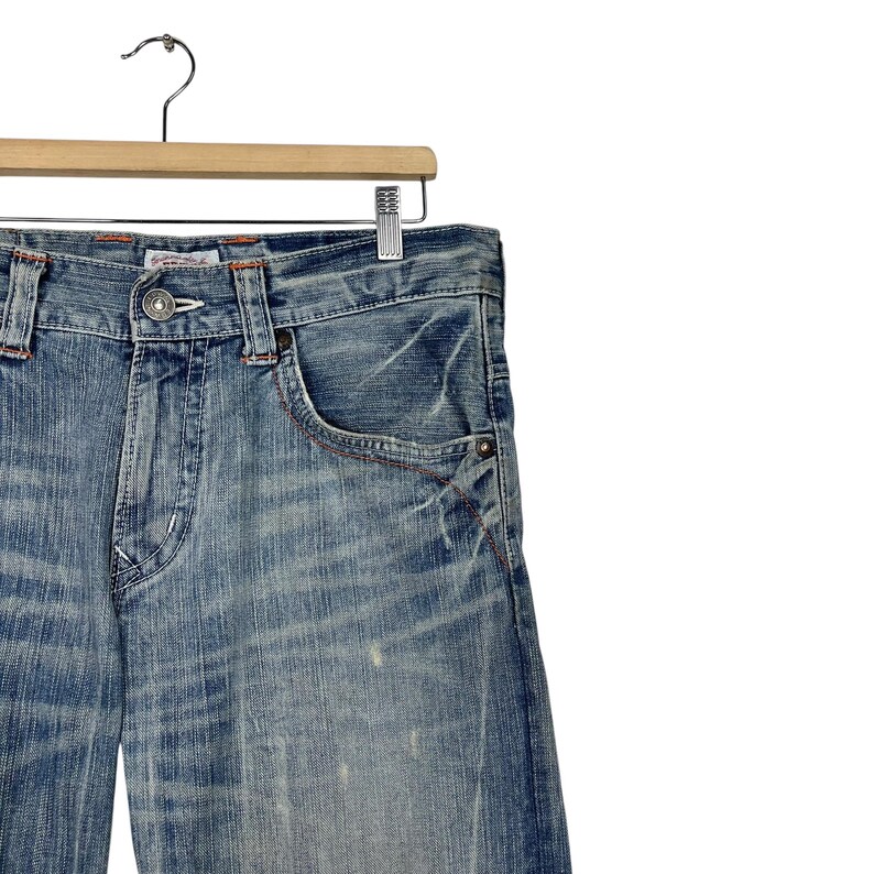 May include: Light blue denim jeans with a faded wash effect, hanging on a wooden hanger. The jeans feature a classic five-pocket design with red stitching accents and a button closure. The fabric shows signs of wear and tear.