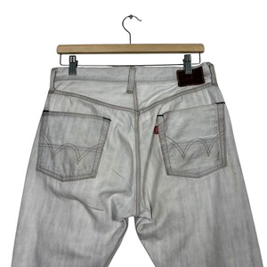 May include: Light gray denim jeans with a classic five-pocket design, visible stitching, and a leather patch. The jeans are displayed on a wooden hanger against a white background.