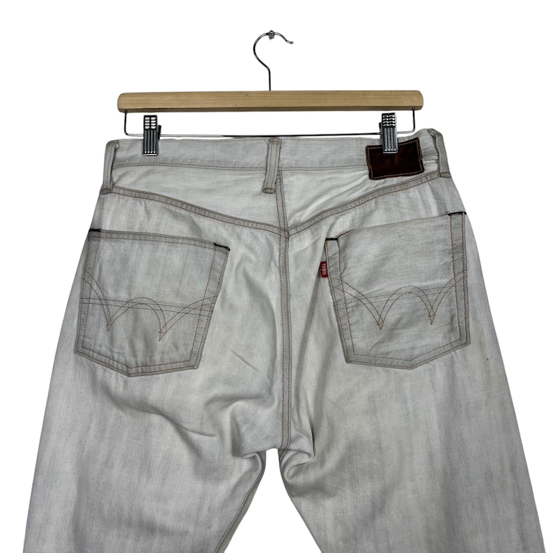May include: Light gray denim jeans with a classic five-pocket design, visible stitching, and a leather patch. The jeans are displayed on a wooden hanger against a white background.