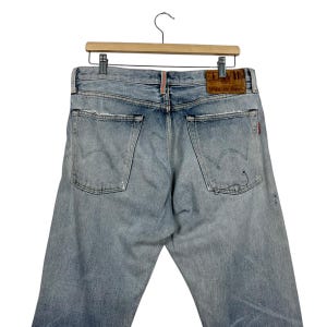 May include: Light blue denim jeans with distressed details, hanging on a wooden hanger. The back pockets feature a stitched design. A leather patch with the brand name is visible.