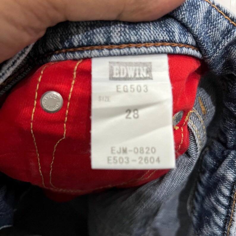 Op de afbeelding: Close-up van de binnenkant van een blauwe spijkerbroek. De rode binnenvoering is zichtbaar, met een wit label met de tekst "EDWIN" en "EG503". Het label toont ook maat "28" en andere productcodes.