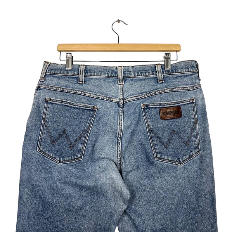 K&ouml;nnte beinhalten: Hellblaue Jeans mit dem Wrangler-Logo auf den Ges&auml;&szlig;taschen. Die Jeans h&auml;ngen an einem Holzb&uuml;gel vor wei&szlig;em Hintergrund. Die Jeans haben ein klassisches F&uuml;nf-Taschen-Design.