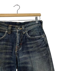 May include: Close-up of blue denim jeans hanging on a wooden hanger. The jeans have a classic five-pocket design, a button and zipper closure, and a faded wash. The jeans are displayed against a white background.