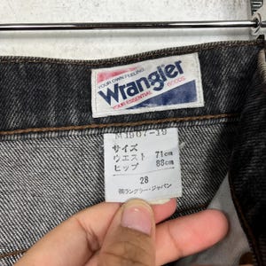 Puede incluir: Primer plano de unos vaqueros Wrangler negros. Los vaqueros tienen una etiqueta con el nombre de la marca y el texto "YOUR OWN FEELING, YOUR ESSENTIAL". La etiqueta incluye informaci&oacute;n de tallas: cintura 71cm, cadera 88cm y talla 28.