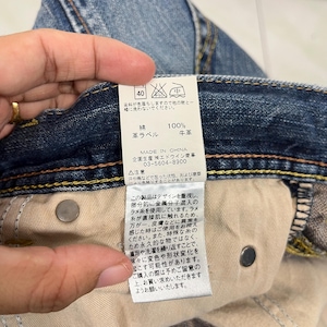 May include: Close-up of a pair of blue denim jeans with a tag attached. The tag has Japanese text and symbols, including washing instructions. The jeans have a button closure and visible stitching.
