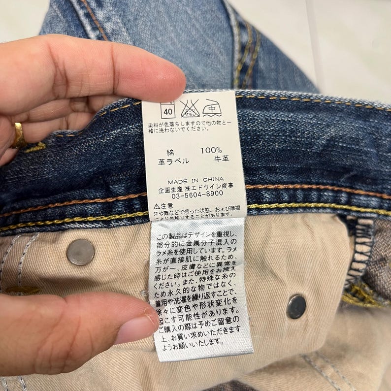 May include: Close-up of a pair of blue denim jeans with a tag attached. The tag has Japanese text and symbols, including washing instructions. The jeans have a button closure and visible stitching.