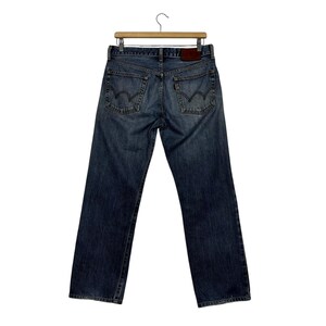 May include: Blue denim jeans with a classic five-pocket design, hanging from a wooden hanger. The jeans have a faded wash, visible stitching, and a leather patch. The back pockets feature a distinctive curved stitch.